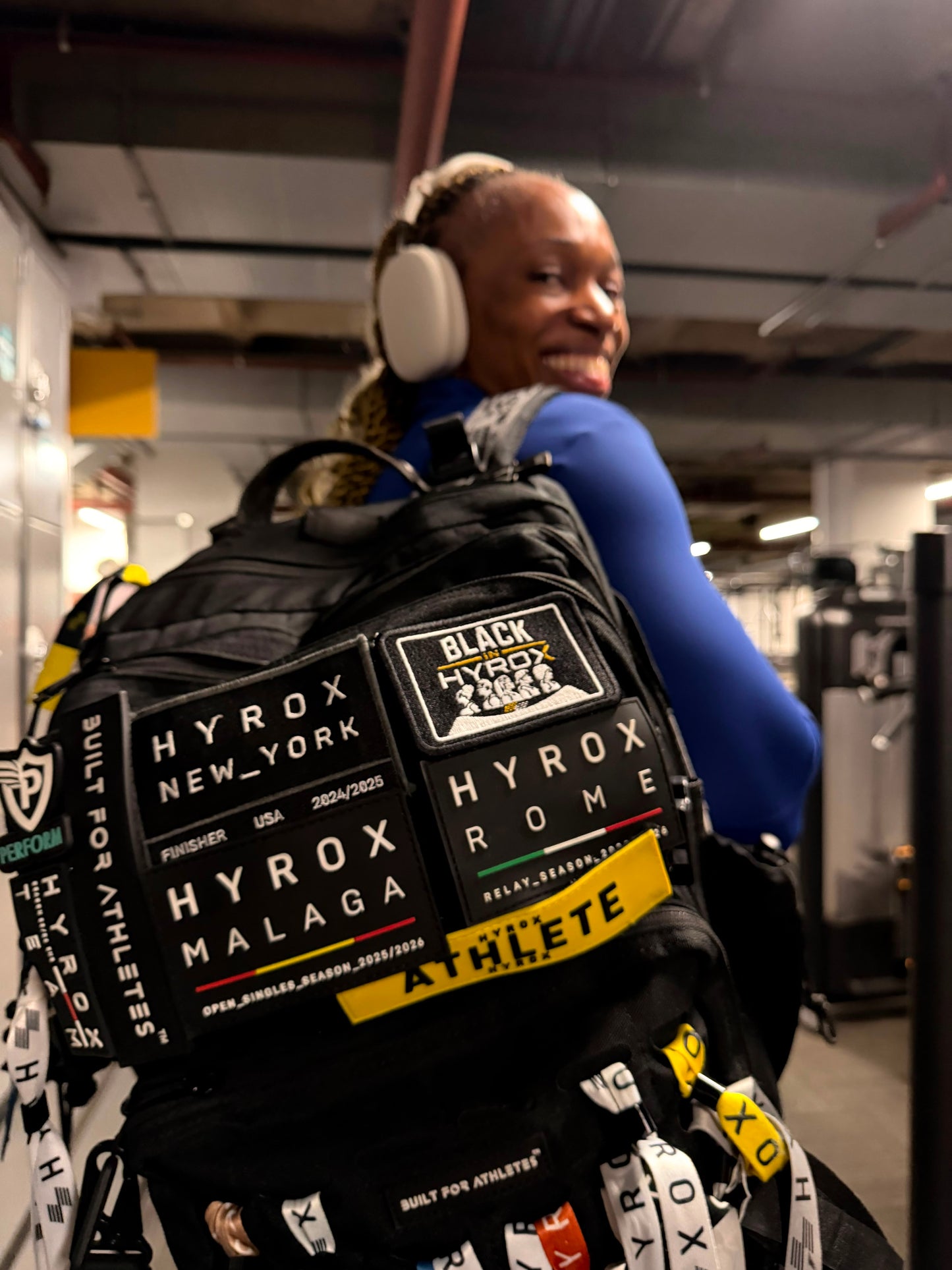 A person in a gym wearing a black Hyrox backpack covered with multiple brand patches and stickers, including Hyrox New York, Hyrox Rome, and Hyrox Malaga.