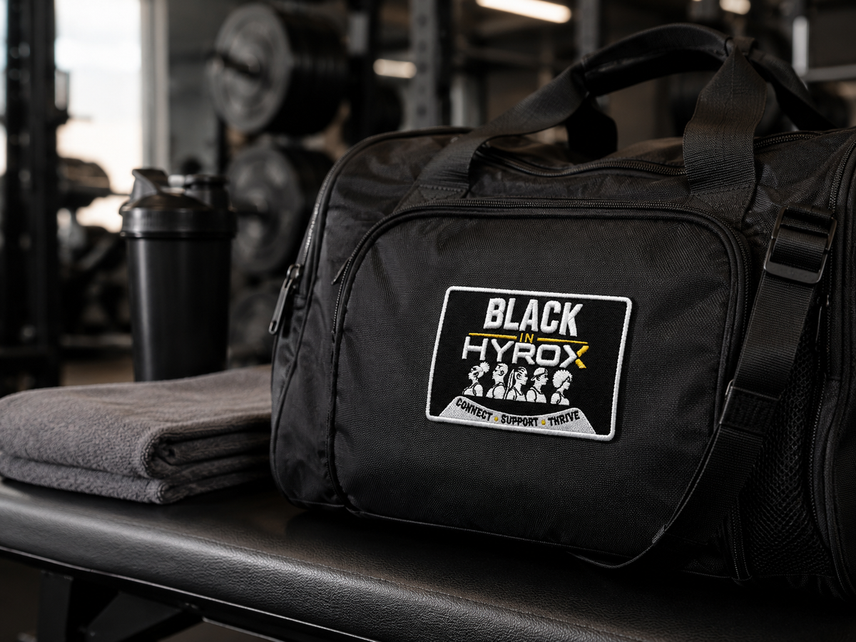 A black gym duffel bag with a 'BLACK in HYROX' patch on the front, resting on a gym bench next to a folded gray towel and a shaker bottle.