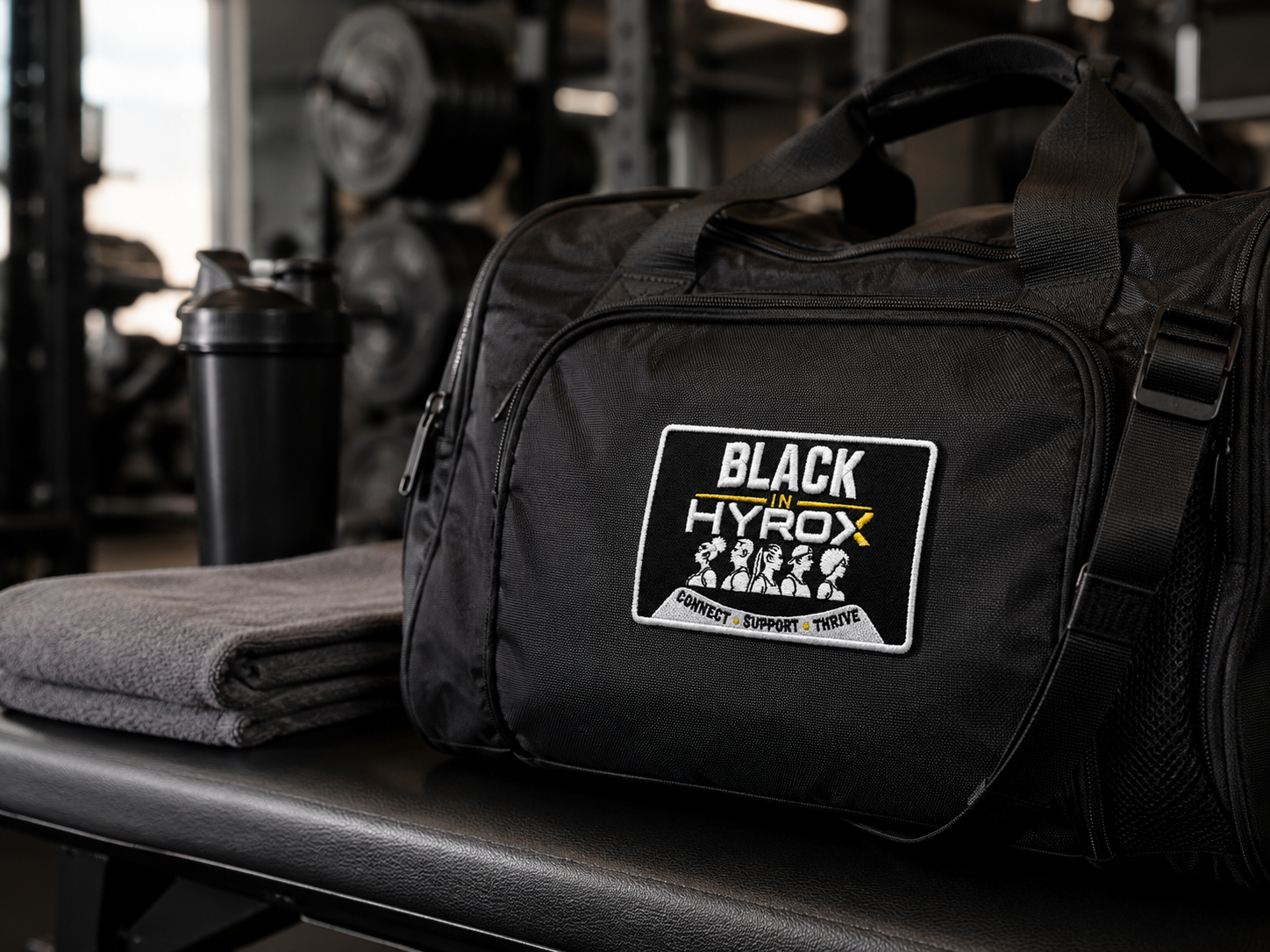 A black gym duffel bag with a 'BLACK in HYROX' patch on the front, resting on a gym bench next to a folded gray towel and a shaker bottle.
