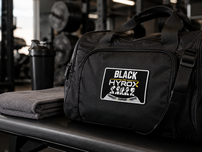 A black gym duffel bag with a 'BLACK in HYROX' patch on the front, resting on a gym bench next to a folded gray towel and a shaker bottle.