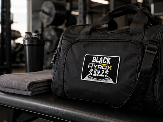 A black gym duffel bag with a 'BLACK in HYROX' patch on the front, resting on a gym bench next to a folded gray towel and a shaker bottle.